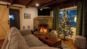 A cozy living room with a stone fireplace, a large window with curtains, a wooden ceiling, and a Christmas tree. - Hot Tub- Newly Renovated Cabin. Modern Rustic (Eureka Springs)