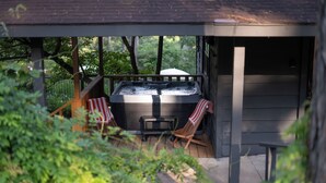 A hot tub on a wooden deck with two striped chairs, surrounded by greenery. - Hot Tub- Newly Renovated Cabin. Modern Rustic (Eureka Springs)