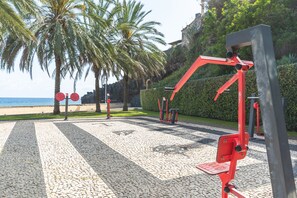 Draußen stehende Trainingsgeräte, Palmen und ein Strand im Hintergrund.