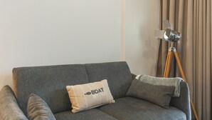Harmaa sohva, jossa on koristetyyny, jonka päällä lukee ”VENE nro 59”, ja jalustalla oleva kamera huoneessa, jossa on beigenväriset verhot.
