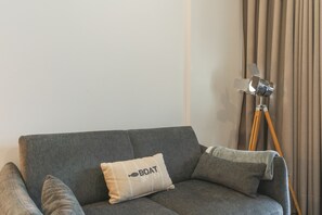 Ein graues Sofa mit einem Kissen mit der Aufschrift 'BOAT No 59' sowie ein Stativ mit einer Kamera in einem Raum mit beigen Vorhängen.