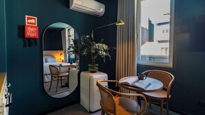 Una camera d'albergo con un letto, un tavolo rotondo con sedie, uno specchio, una pianta in vaso e una finestra con tende.