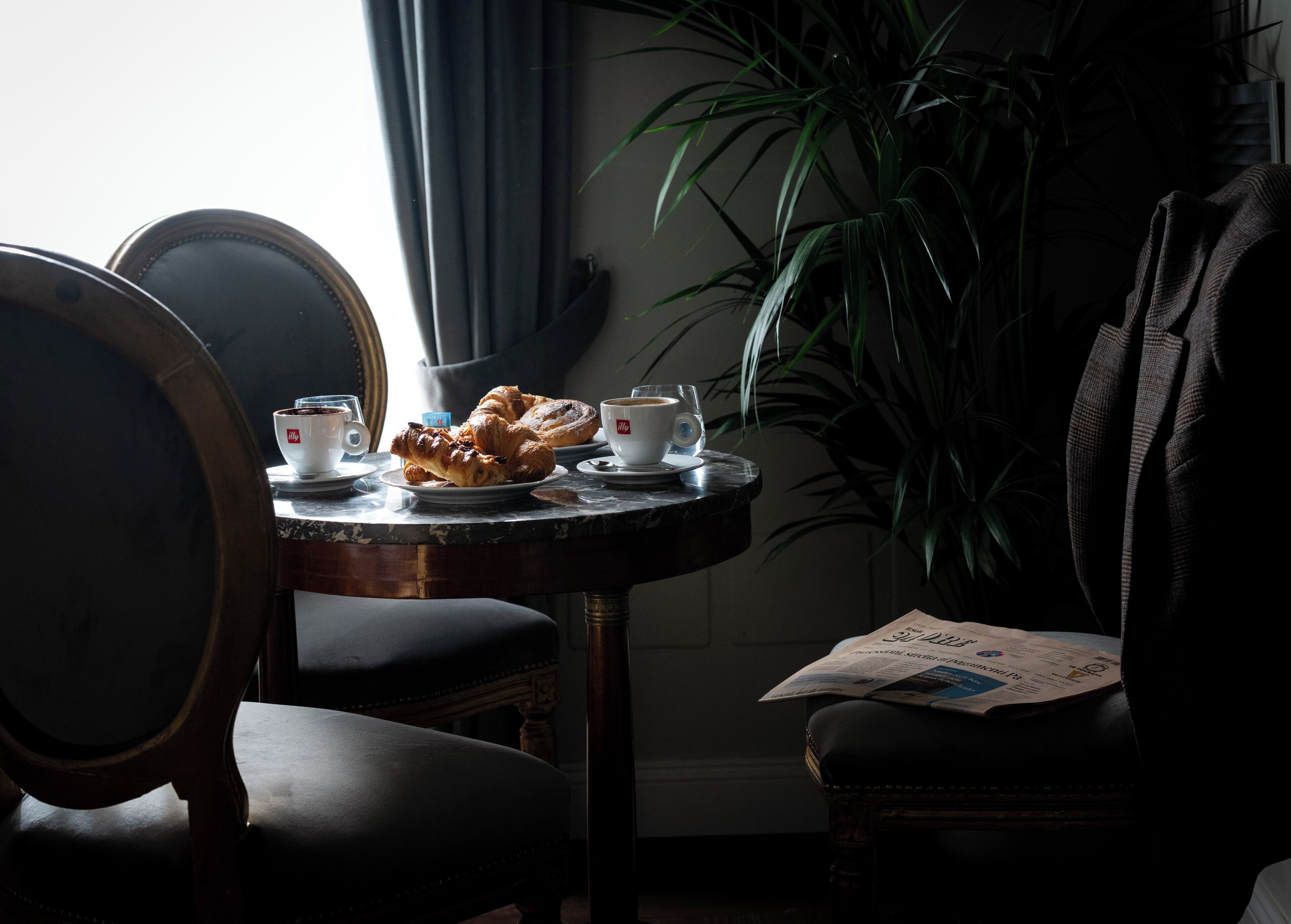 A dining table with croissants, coffee cups, and a menu. There are chairs around the table, and a plant is in the background.