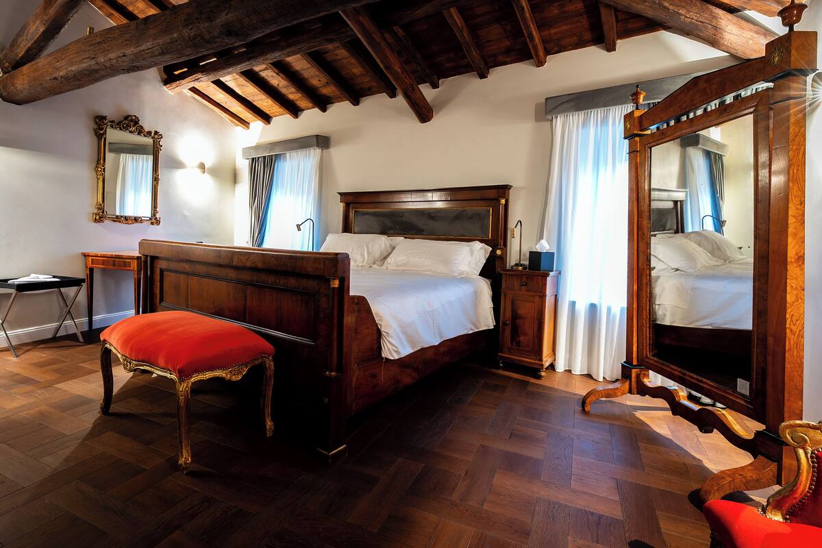 A bedroom with a wooden bed, a red upholstered bench, a wooden nightstand, a mirror, and a wooden chair.