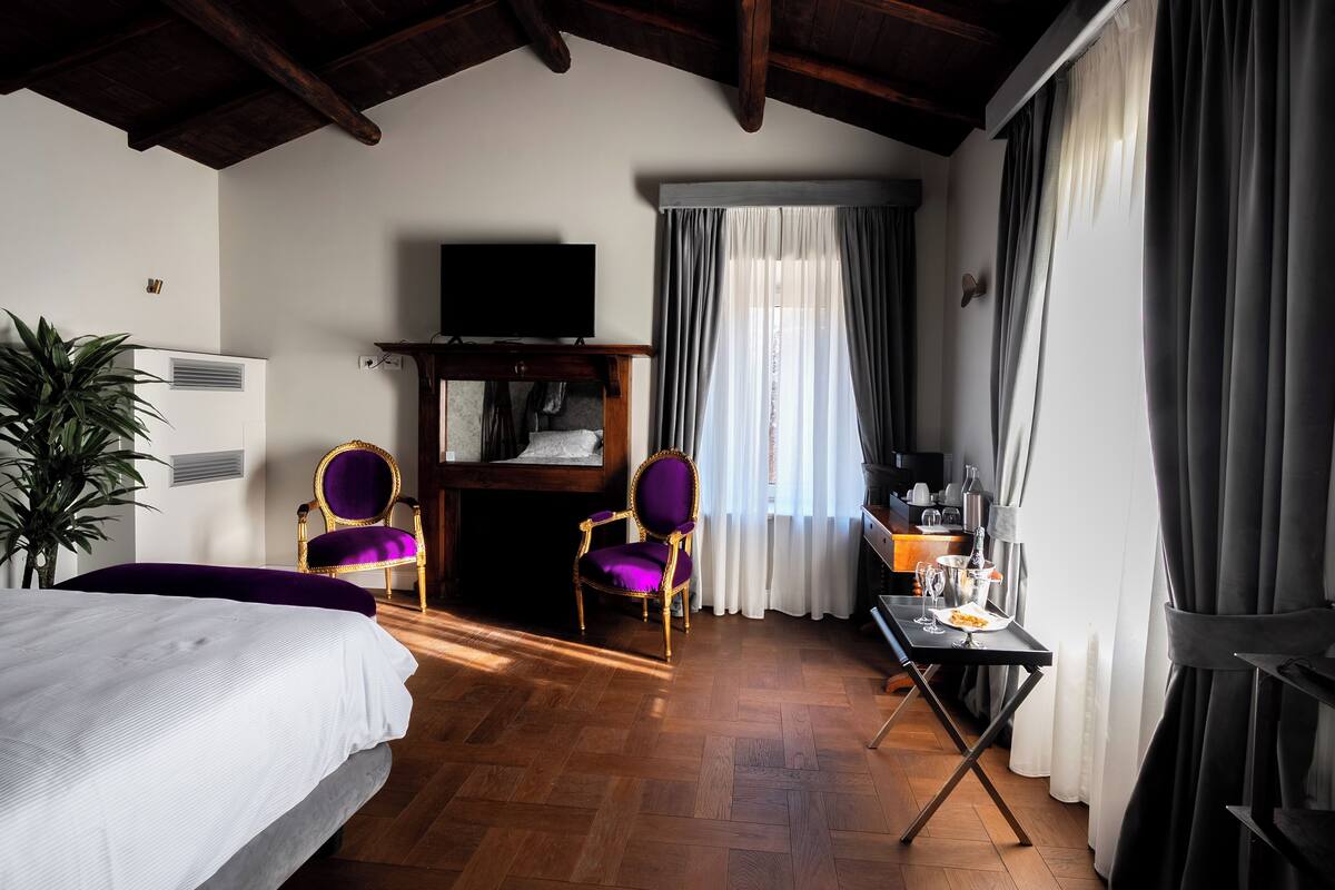 A hotel room with a bed, two purple armchairs, a small table with a plate of food, a television, and a window with curtains.