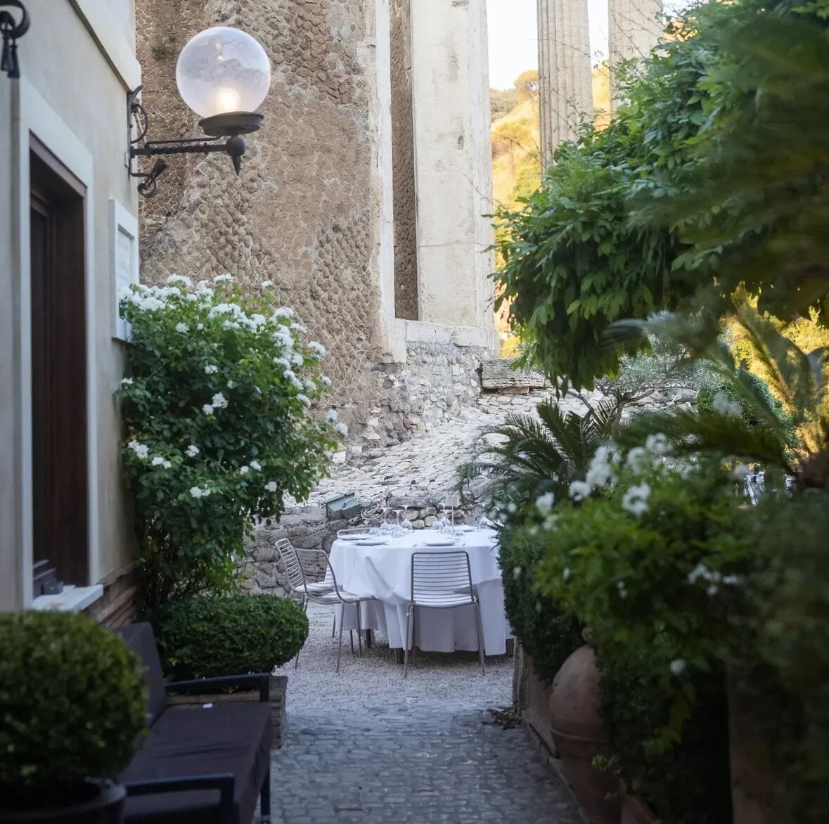 An outdoor dining area with tables and chairs, surrounded by greenery and stone walls.