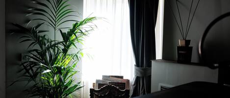 A room with a potted plant, a window with sheer curtains, a small table with books, and a wall-mounted shelf with a candle and reed diffuser.