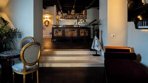 Un área de bar con barra de madera, estantes con botellas y un piano. Hay sillas y una mesa con una lámpara.