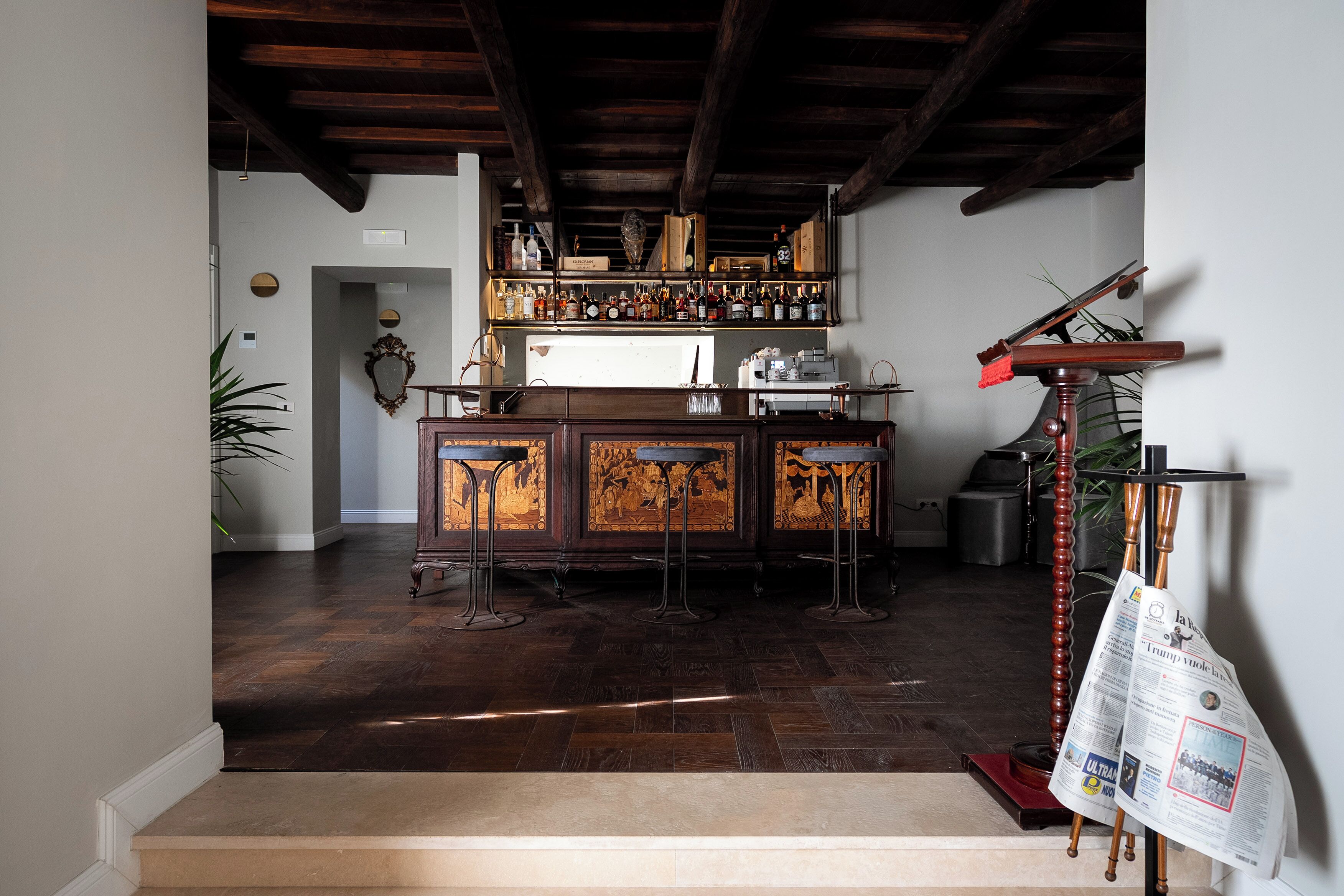 A bar area with a wooden bar counter, stools, and a shelf with bottles.