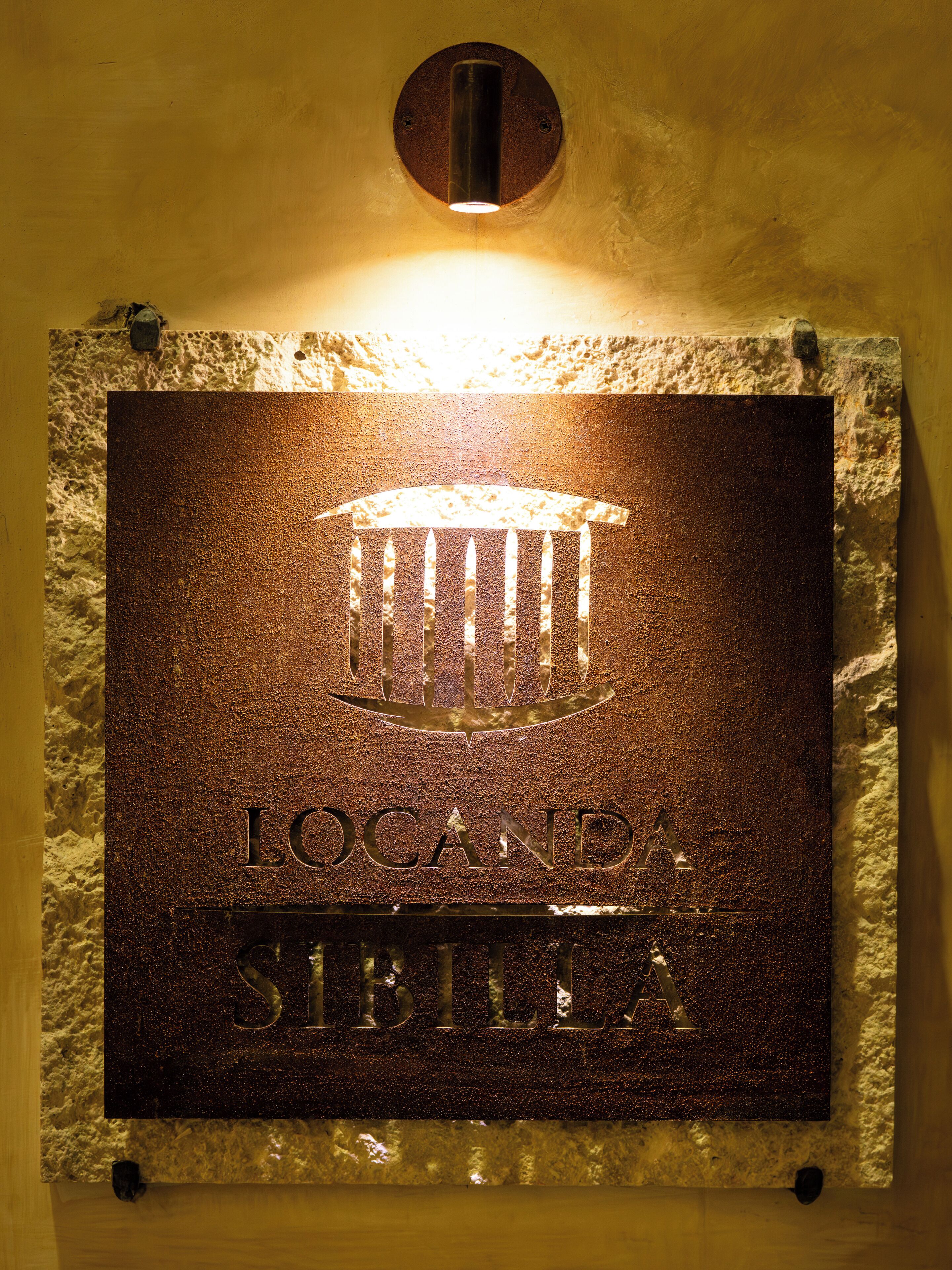 A plaque with the inscription "LOCANDA SIBILLA" mounted on a textured wall, illuminated by a light fixture above.