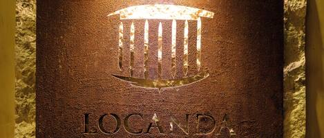 A plaque with the inscription "LOCANDA SIBILLA" mounted on a textured wall, illuminated by a light fixture above.