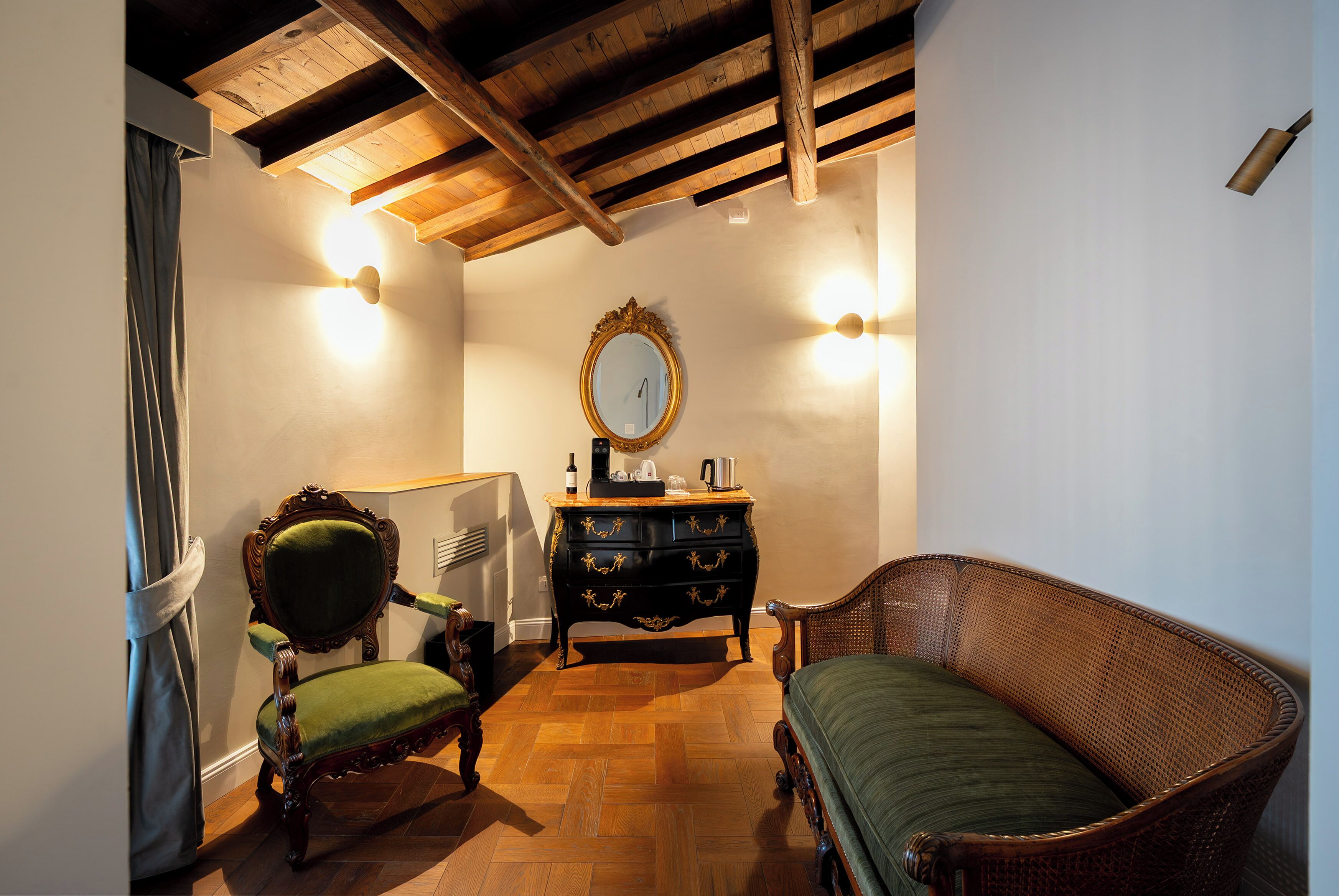 Ein Zimmer mit Holzboden, einem grünen Polsterstuhl, einem hölzernen Sofa, einer dunklen Holzkommode mit Goldakzenten und einem Spiegel an der Wand.
