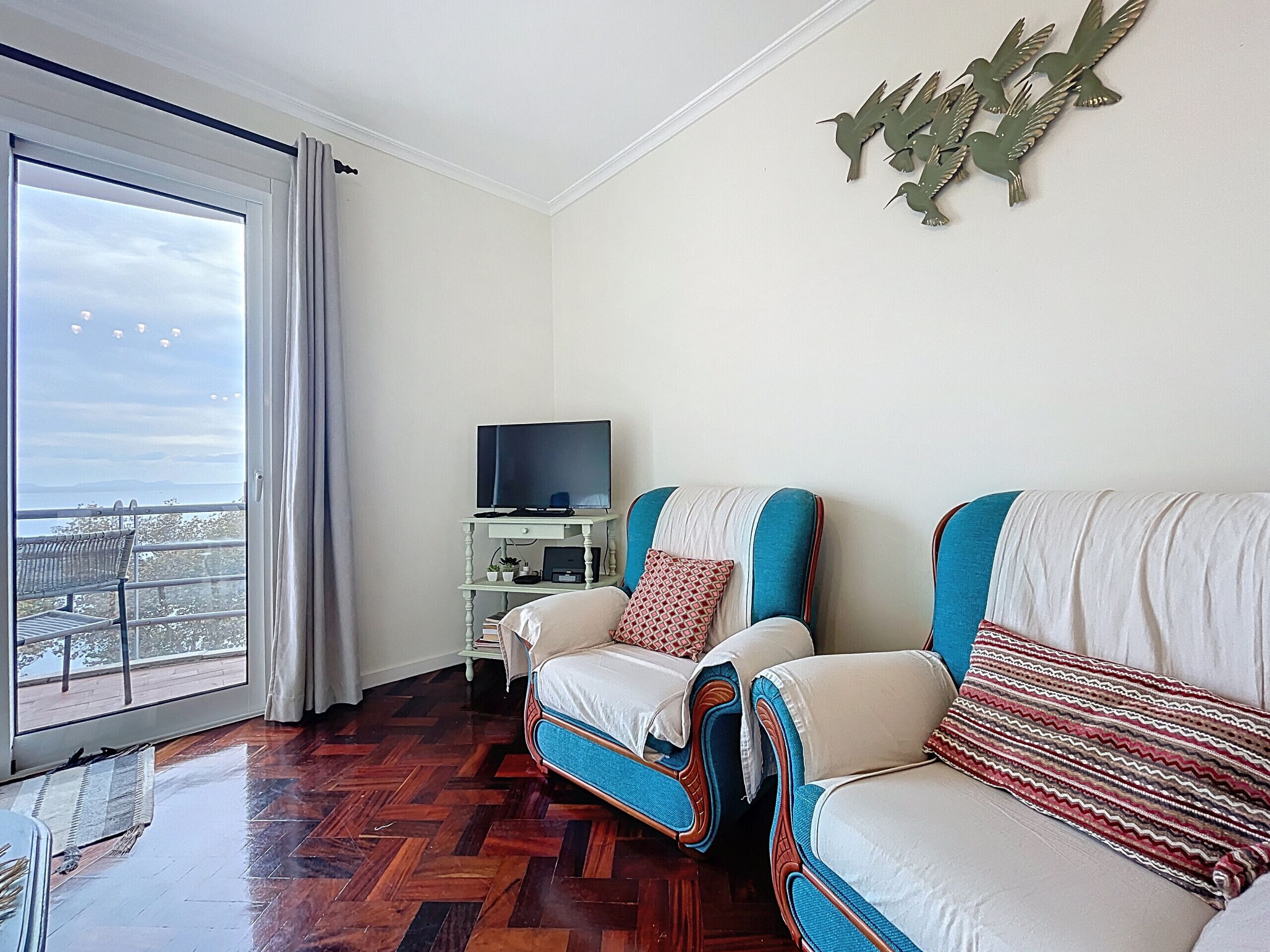 A room with a large window, a flat-screen TV on a small table, two sofas with patterned cushions, and a decorative wall piece.