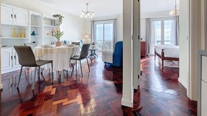 Ein geräumiges Zimmer mit Holzboden im Fischgrätmuster, einem Essbereich mit weißem Tisch und Stühlen sowie einem großen Fenster mit Vorhängen.