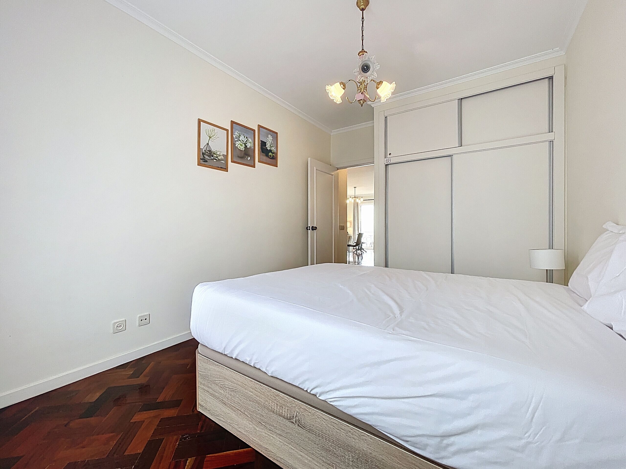 A bedroom with a large bed, a chandelier, a door leading to another room, and three framed pictures on the wall.