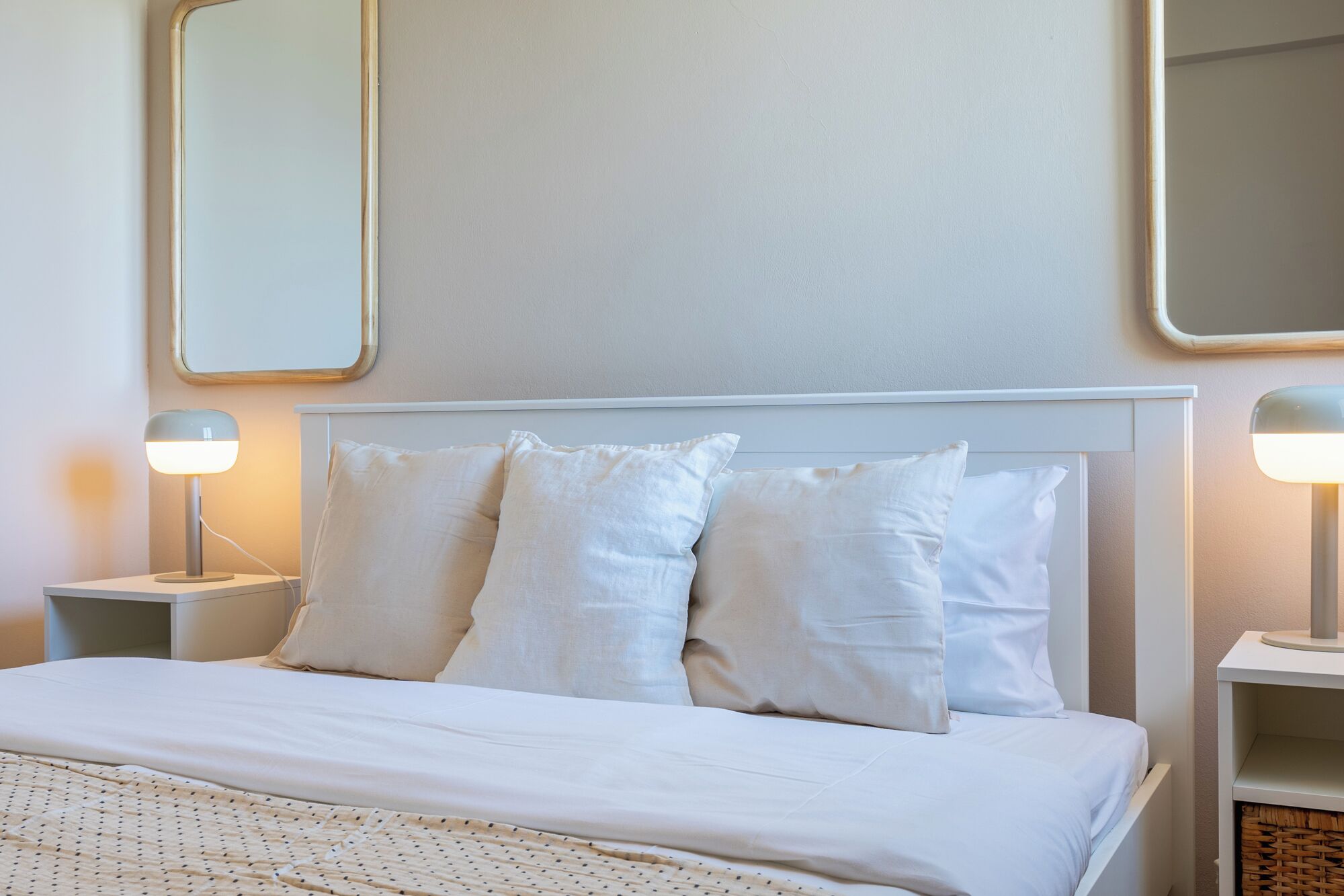 Ein ordentlich bezogenes Bett mit weißen Leinen und Kissen, seitlich flankiert von zwei Nachttischen mit Lampen, in einem Zimmer mit schlichten Wänden und einem großen Spiegel über dem Bett.
