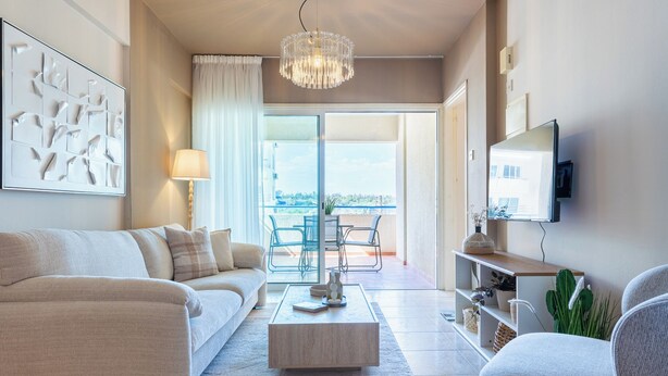 A modern living room with a sofa, coffee table, and chairs. There is a balcony with a view, a chandelier, and a TV mounted on the wall. - Calina - 3-BR Apt & Pool in Mackenzie (Larnaca)