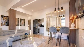 A modern living room with a sofa, a coffee table, and two bar stools. There is a kitchen in the background with a dining area. - Golden Hour Residence - Elegant Cityview Apartment in Historic Bucharest (București)