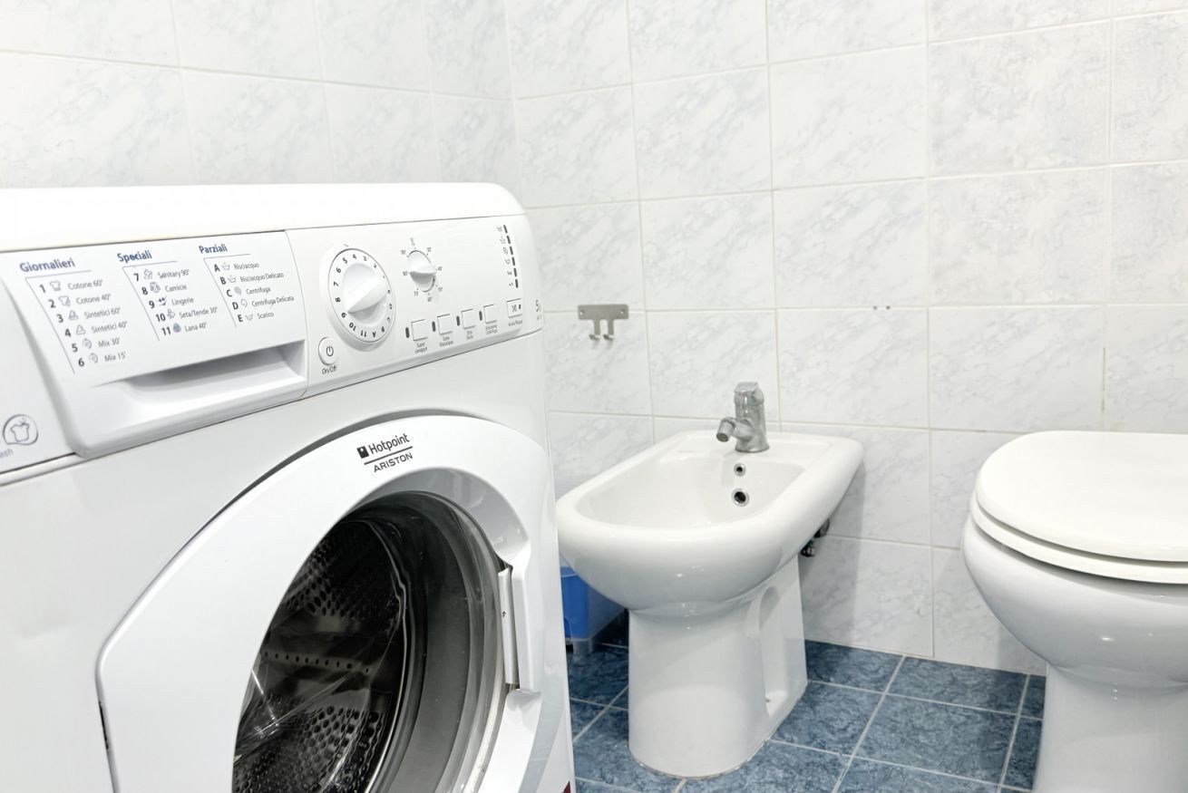 Een wasmachine met een voorlaadopening, een bidet en een toilet in een witte betegelde badkamer.