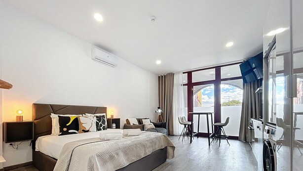Una habitación de hotel moderna con una cama grande, un escritorio con dos sillas, vista a la ciudad y un balcón con vista al cielo.