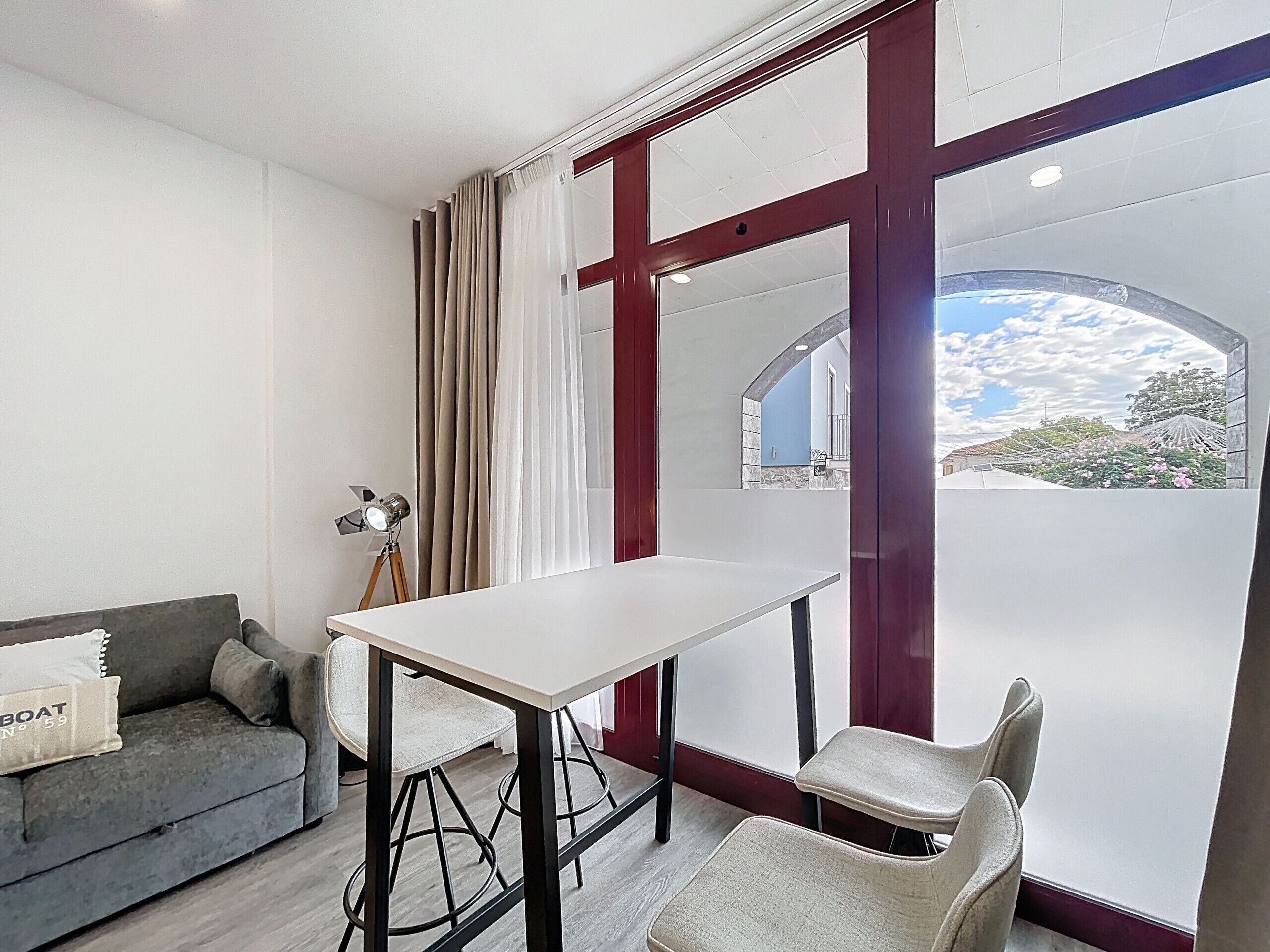 Ein modernes Zimmer mit einem weißen Tisch und zwei Stühlen, einem grauen Sofa und einem großen Fenster mit Blick auf den Himmel und die Gebäude.
