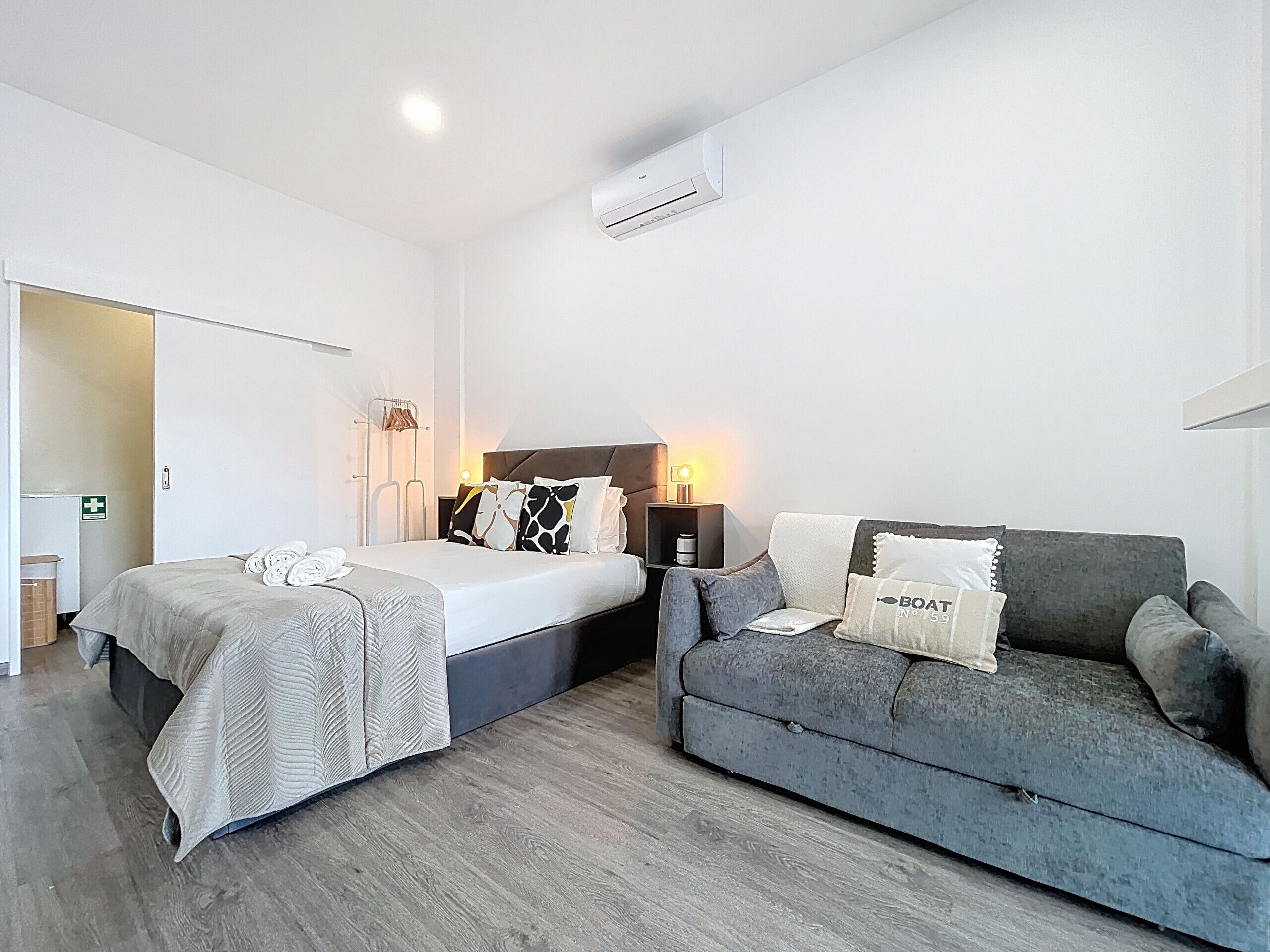 A hotel room with two beds, a sofa, a bedside table with a lamp, and an air conditioning unit.