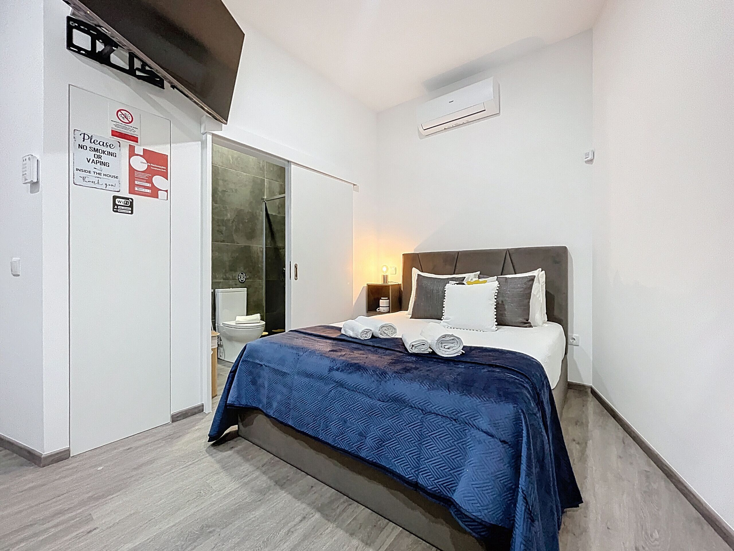 Ein Schlafzimmer mit Bett, Nachttisch, Wand-Klimaanlage und einer Tür mit Rauchverbot-Schild.