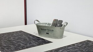 Ein Metallbehälter mit dem Aufdruck 'Garden Edition Vintage Container', gefüllt mit Fernbedienungen, steht auf einem weißen Tisch mit gemusterten Untersetzern.