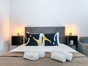 Ein ordentlich bezogenes Bett mit grauem Kopfteil, zwei Nachttische mit Lampen und zusammengerollte Handtücher auf dem Bett.