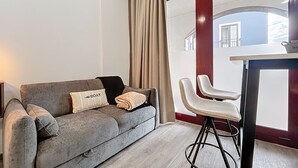 Ein modernes Wohnzimmer mit einem grauen Sofa, zwei Sesseln, einem kleinen Tisch und einem großen Fenster mit Vorhängen.