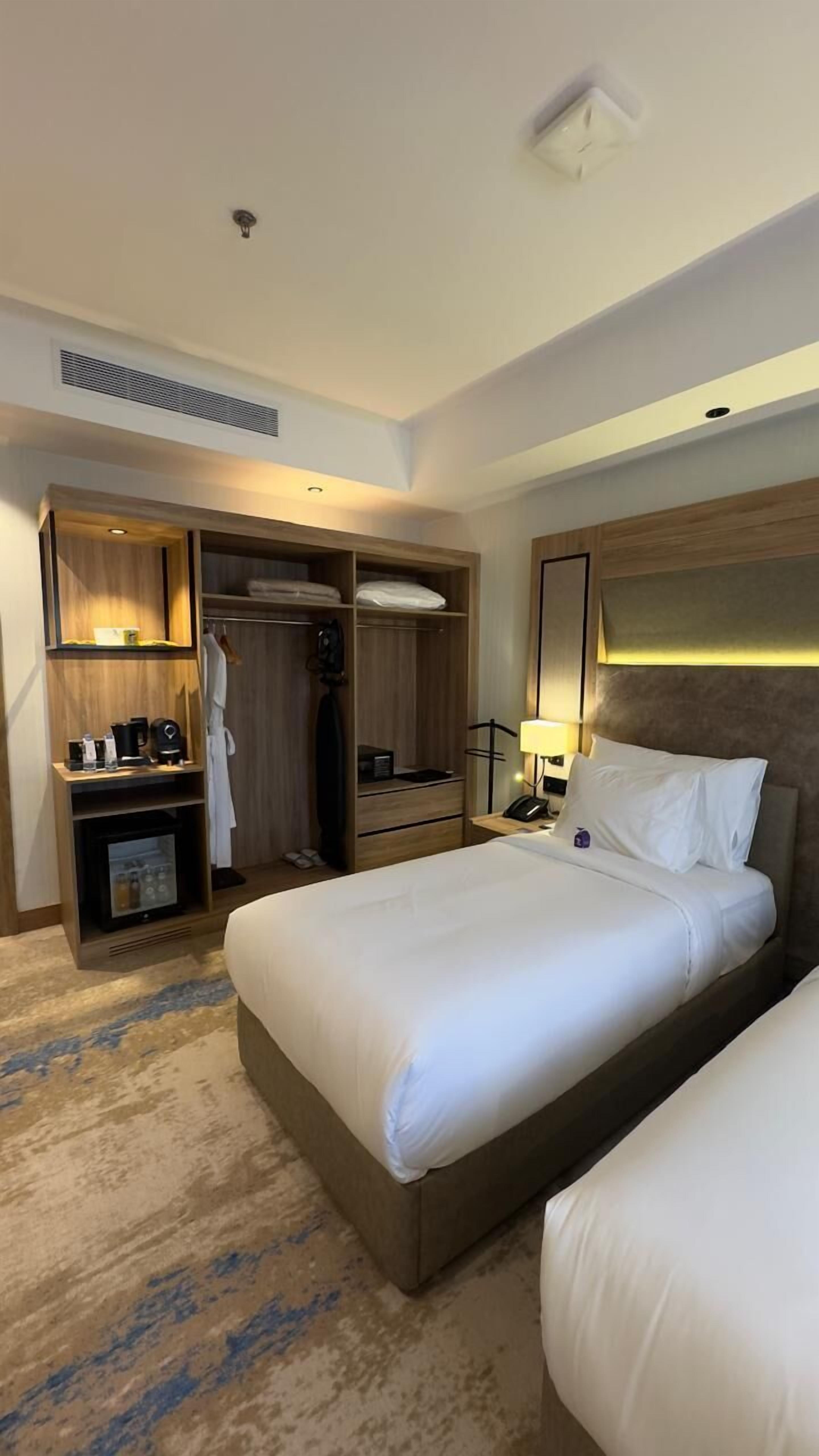 A hotel room with a large bed, a wardrobe, a desk with a lamp, and a minibar.
