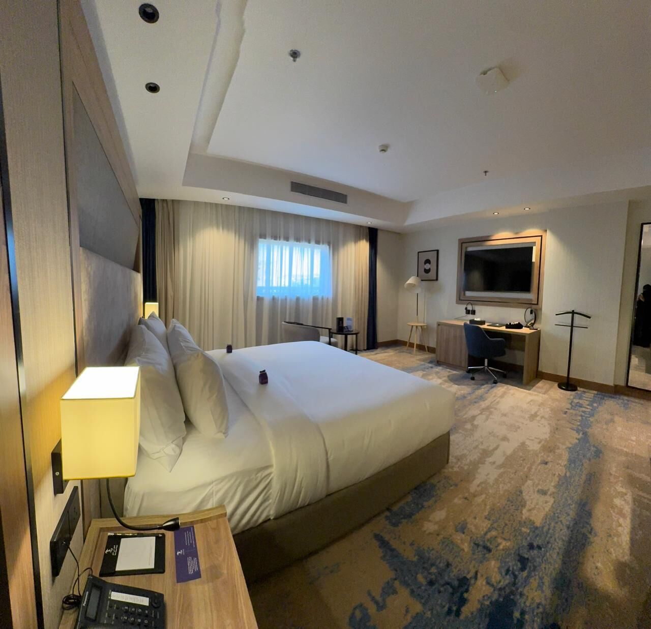 A hotel room with a large bed, a desk, a TV, and a window with curtains.