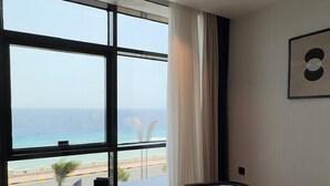 Ένα σύγχρονο δωμάτιο ξενοδοχείου με ένα μεγάλο παράθυρο που προσφέρει θέα στον ωκεανό. Υπάρχει ένα στρογγυλό τραπέζι με μια καρέκλα, ένας πίνακας σε κορνίζα στον τοίχο και ένα μικρό βοηθητικό τραπεζάκι με ένα φωτιστικό.