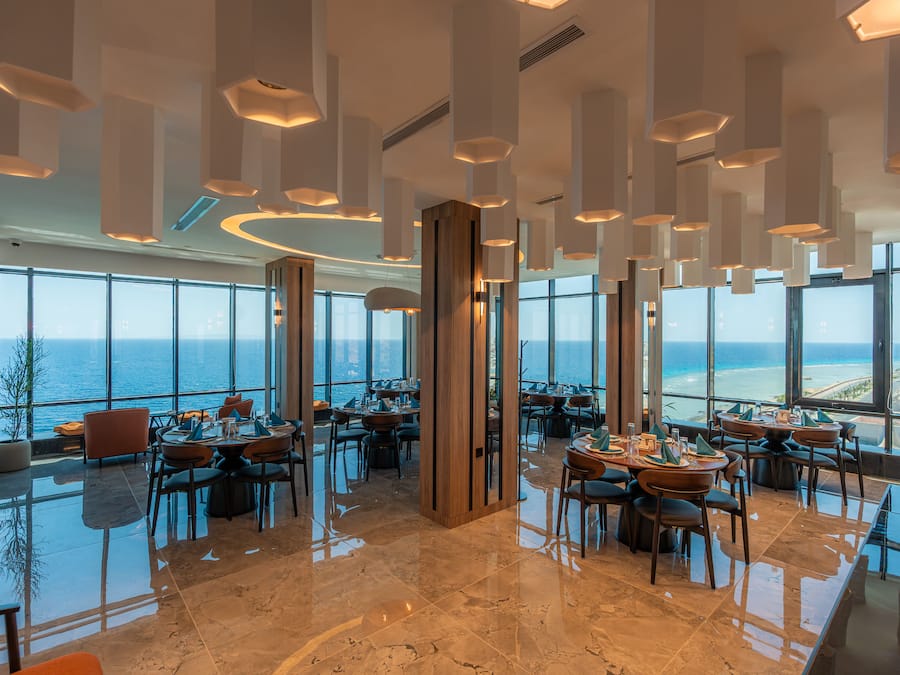 A modern restaurant with a panoramic ocean view, featuring high ceilings, contemporary lighting, and a polished marble floor.