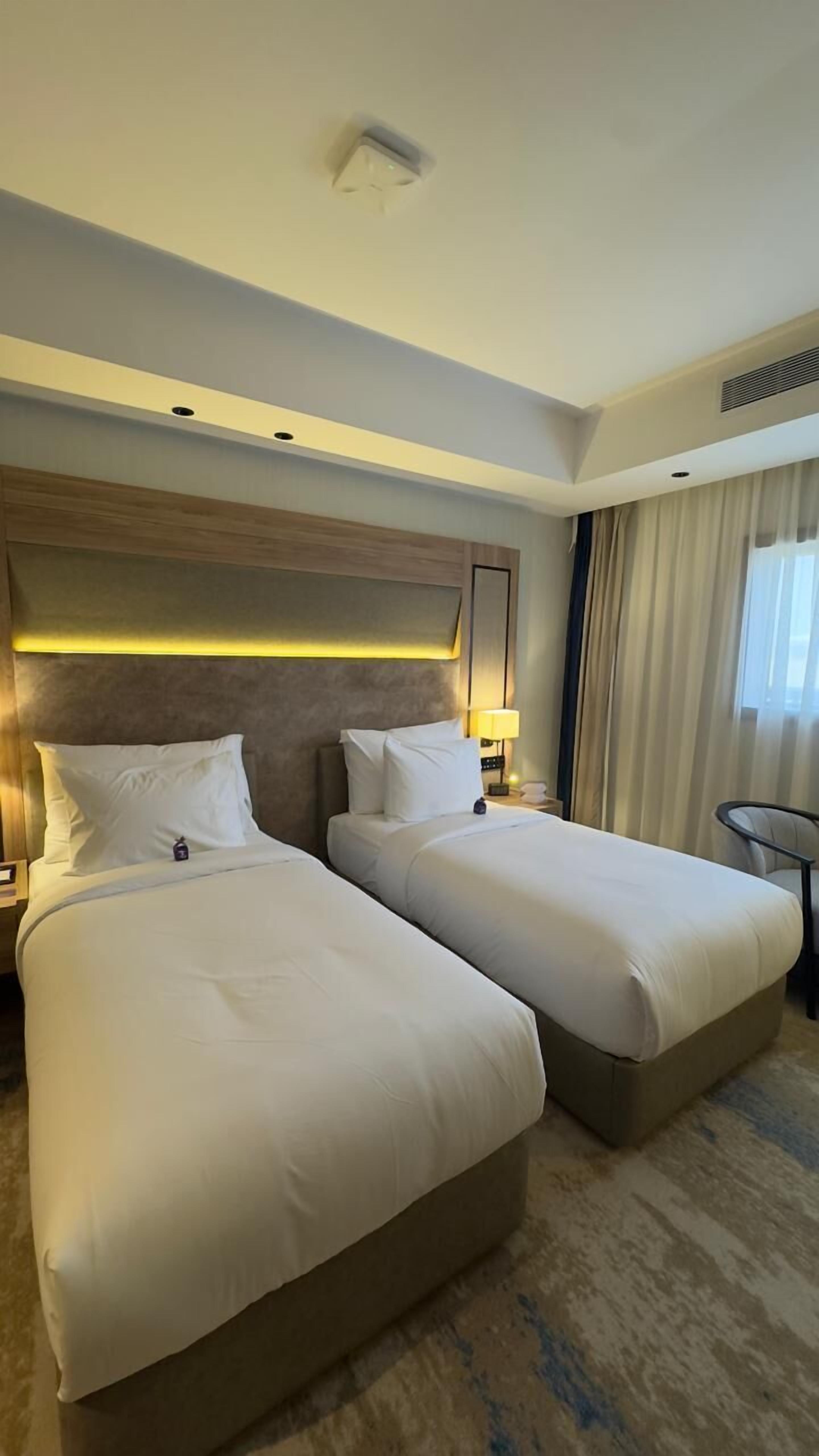 A hotel room with two beds, a headboard with integrated lighting, a bedside table with a lamp, and a view of the outdoors through a window with curtains.