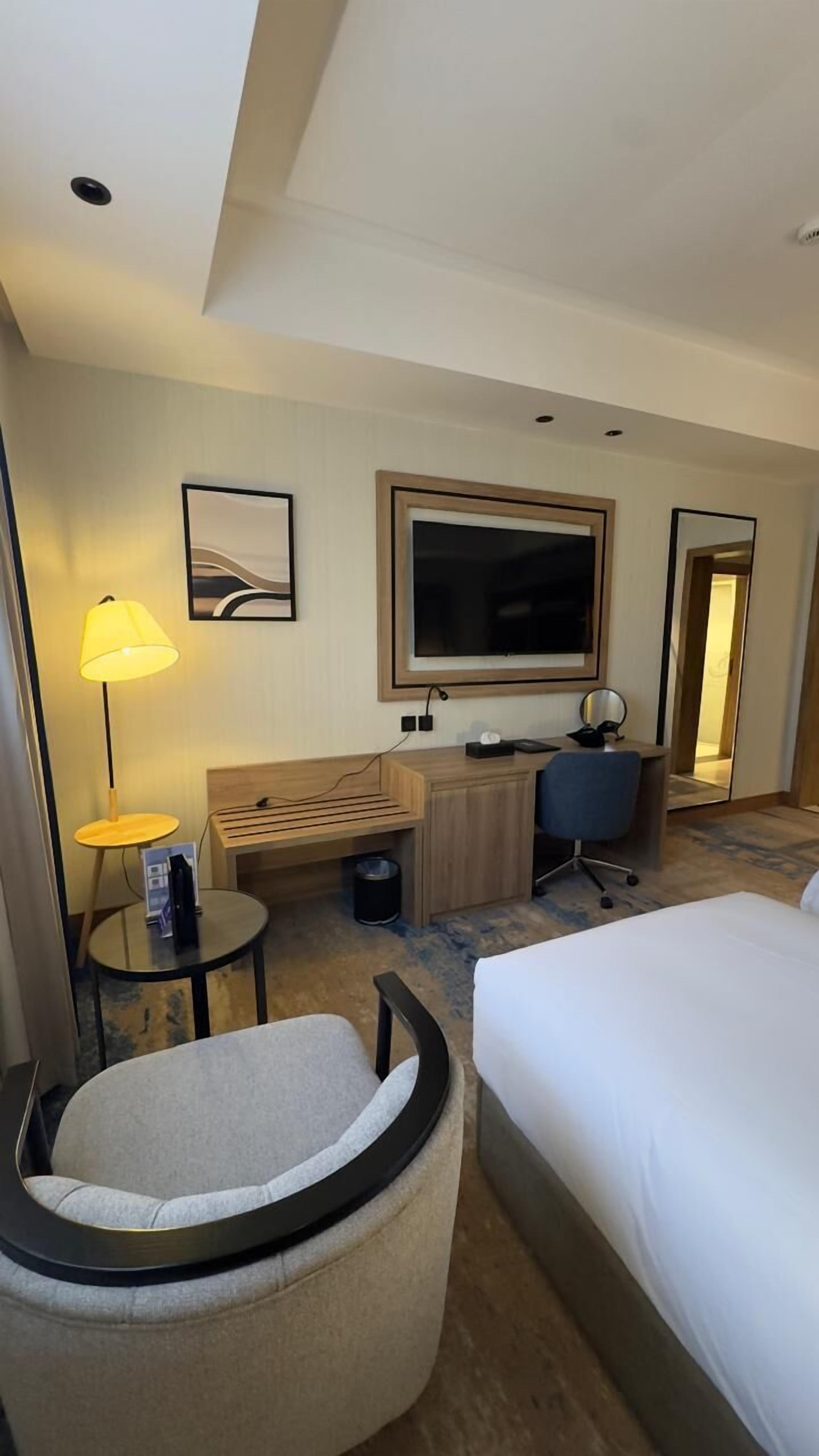 A hotel room with a bed, a desk, a chair, a TV, and a lamp.