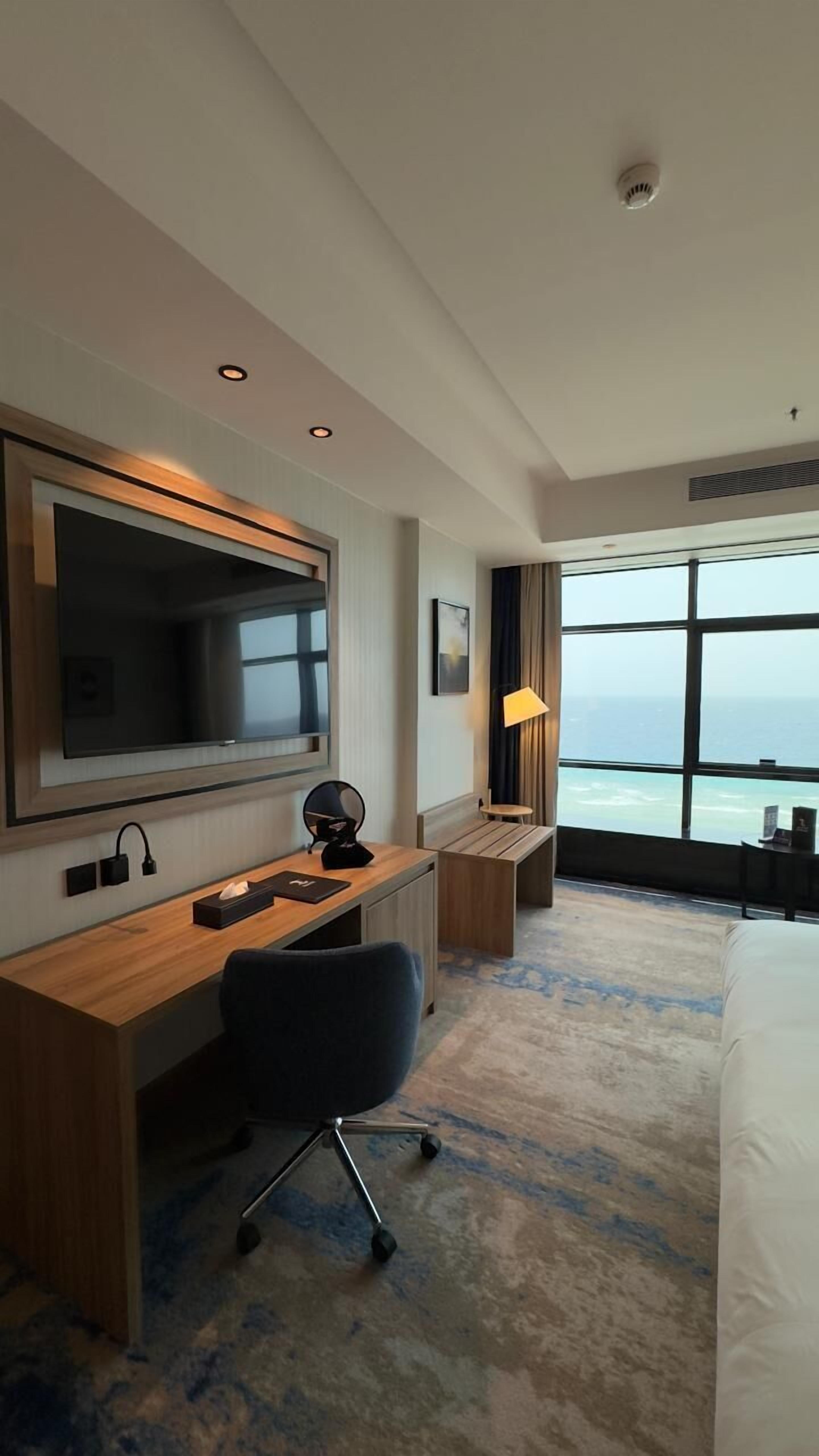A hotel room with a large TV, a desk with a chair, a lamp, and a view of the ocean.