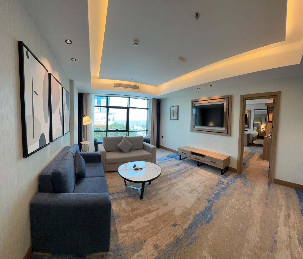 Una habitación de hotel moderna con sofá, mesa de centro, televisión y un amplio ventanal con vistas a la ciudad.