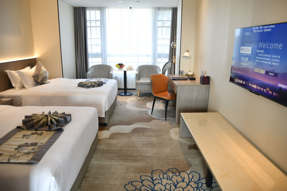 A hotel room with two beds, a desk, a chair, a TV, and a view of the outdoors.