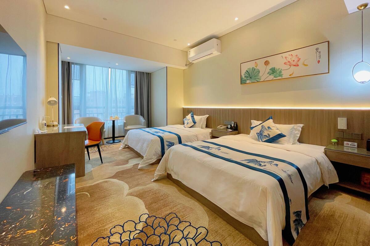 A hotel room with two beds, a desk, a chair, a small table, an air conditioning unit, and a wall-mounted artwork.