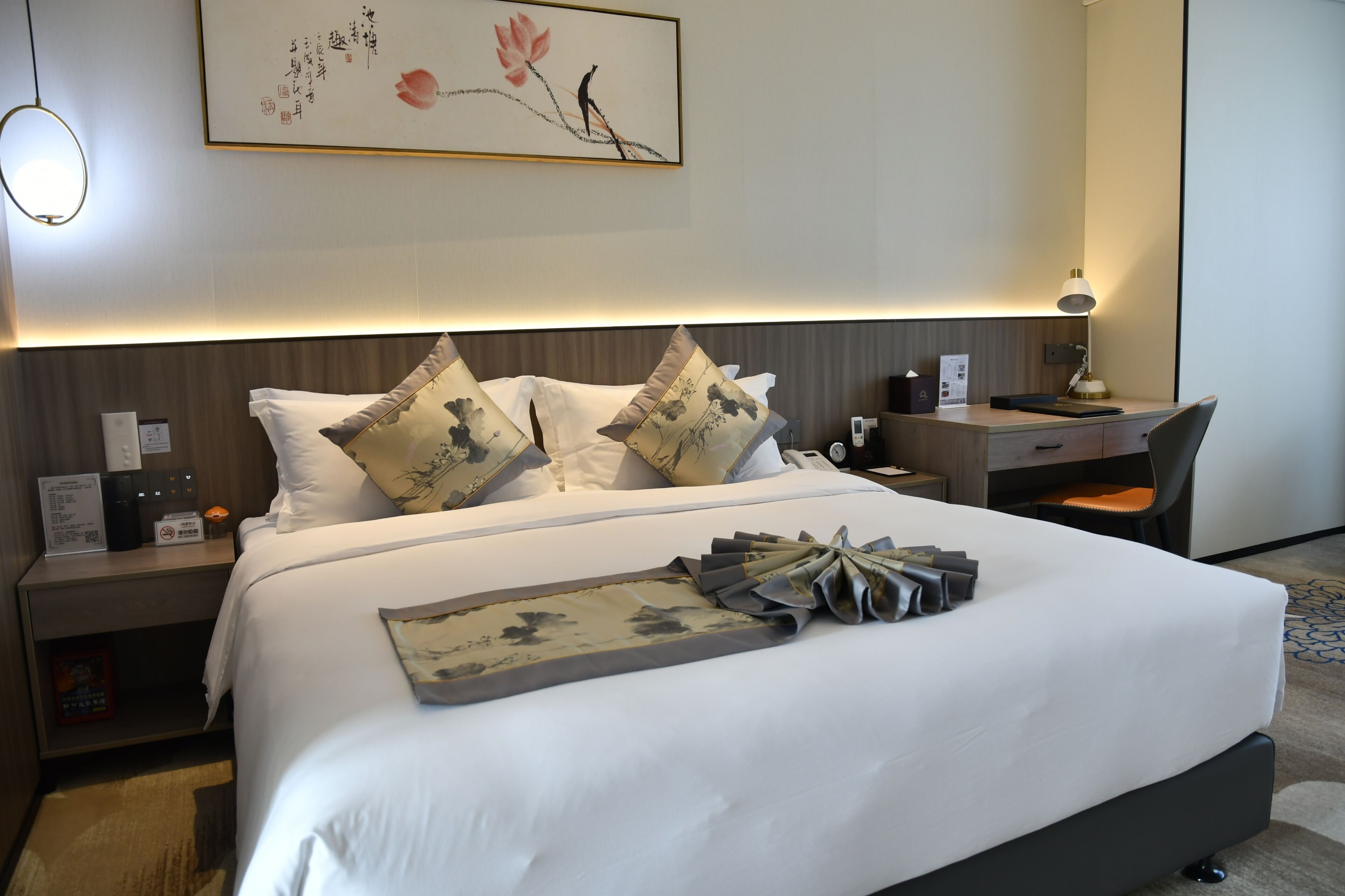 Ein Hotelzimmer mit einem ordentlich bezogenen Bett, einem Schreibtisch und einem Stuhl. An der Wand hängt ein Gemälde, auf dem Schreibtisch eine Lampe und auf dem Nachttisch ein Telefon.