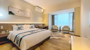 Een moderne hotelkamer met een groot bed, een bureau, twee fauteuils, een bijzettafeltje en een groot raam met gordijnen.