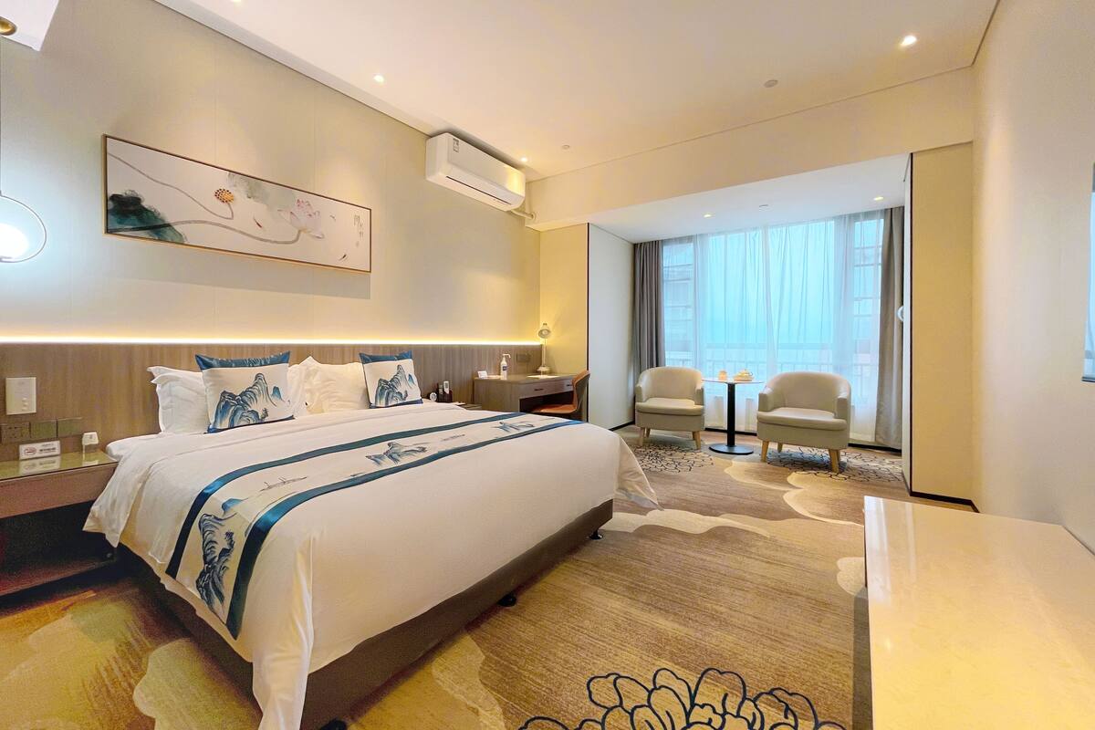 A modern hotel room with a large bed, a desk, two armchairs, a small table, and a large window with curtains.