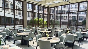 Een ruime eetruimte met meerdere ronde tafels en stoelen, grote ramen met uitzicht op gebouwen en groen.