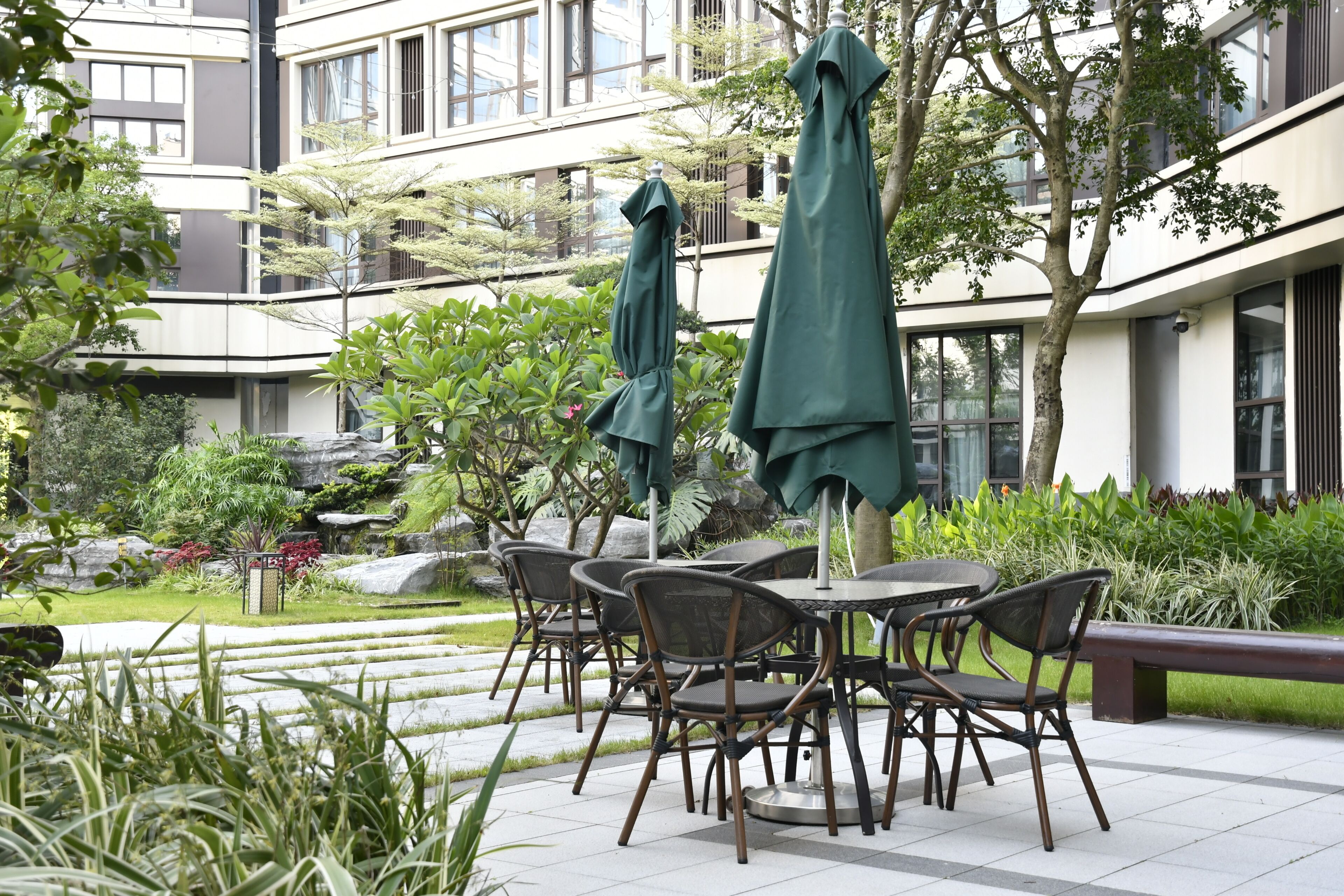 An outdoor seating area with tables and chairs, surrounded by greenery and a building in the background.