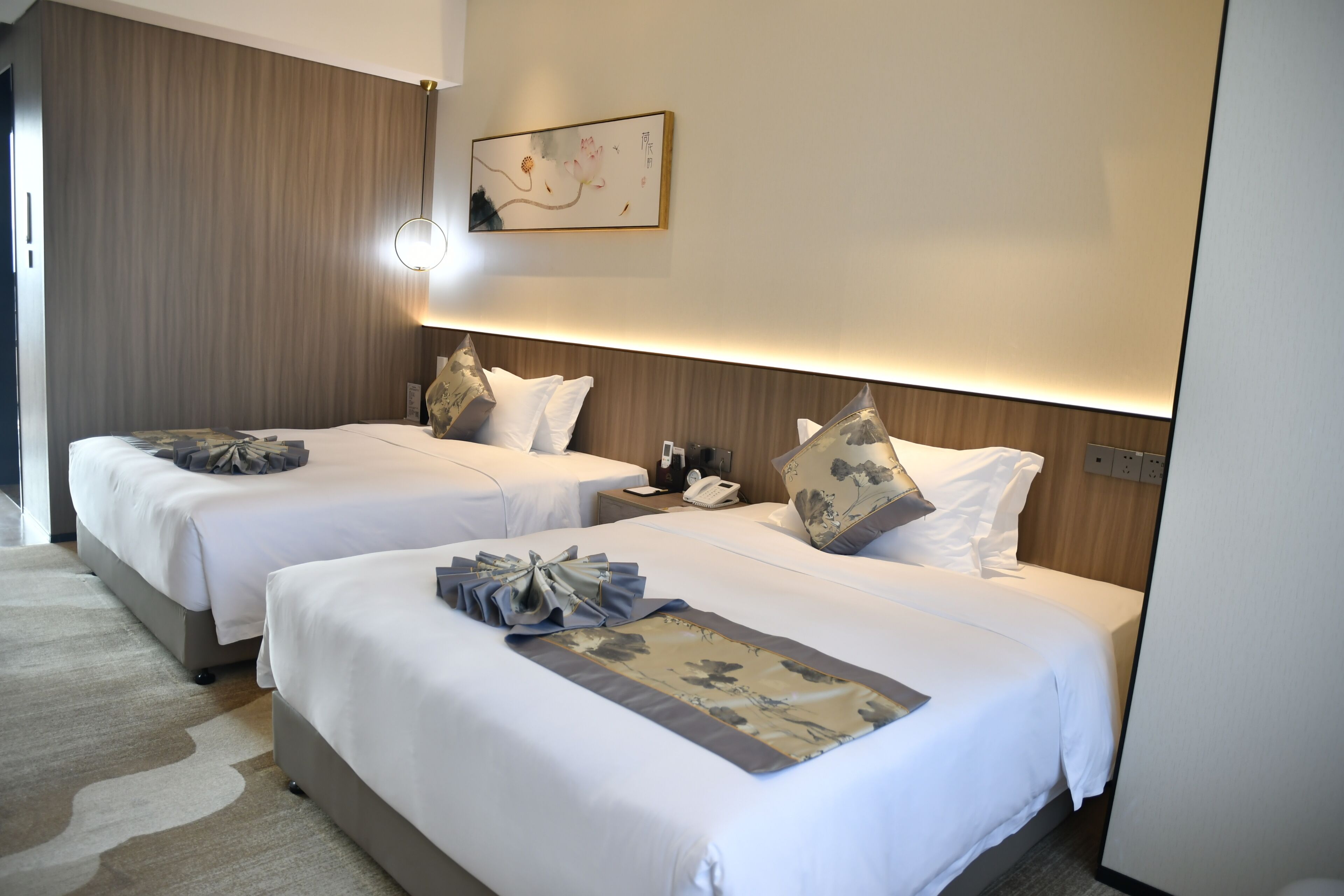 A hotel room with two beds, a bedside table with a lamp, a painting on the wall, and a carpeted floor.