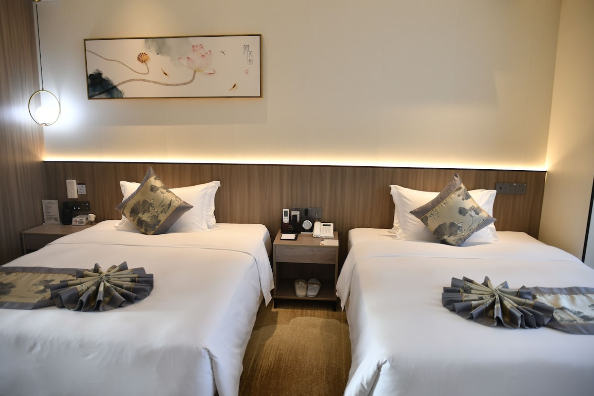 A hotel room with two beds, each with a decorative pillow and a folded fabric on the bed. There is a nightstand with a phone and a clock, and a wall-mounted artwork above the beds.