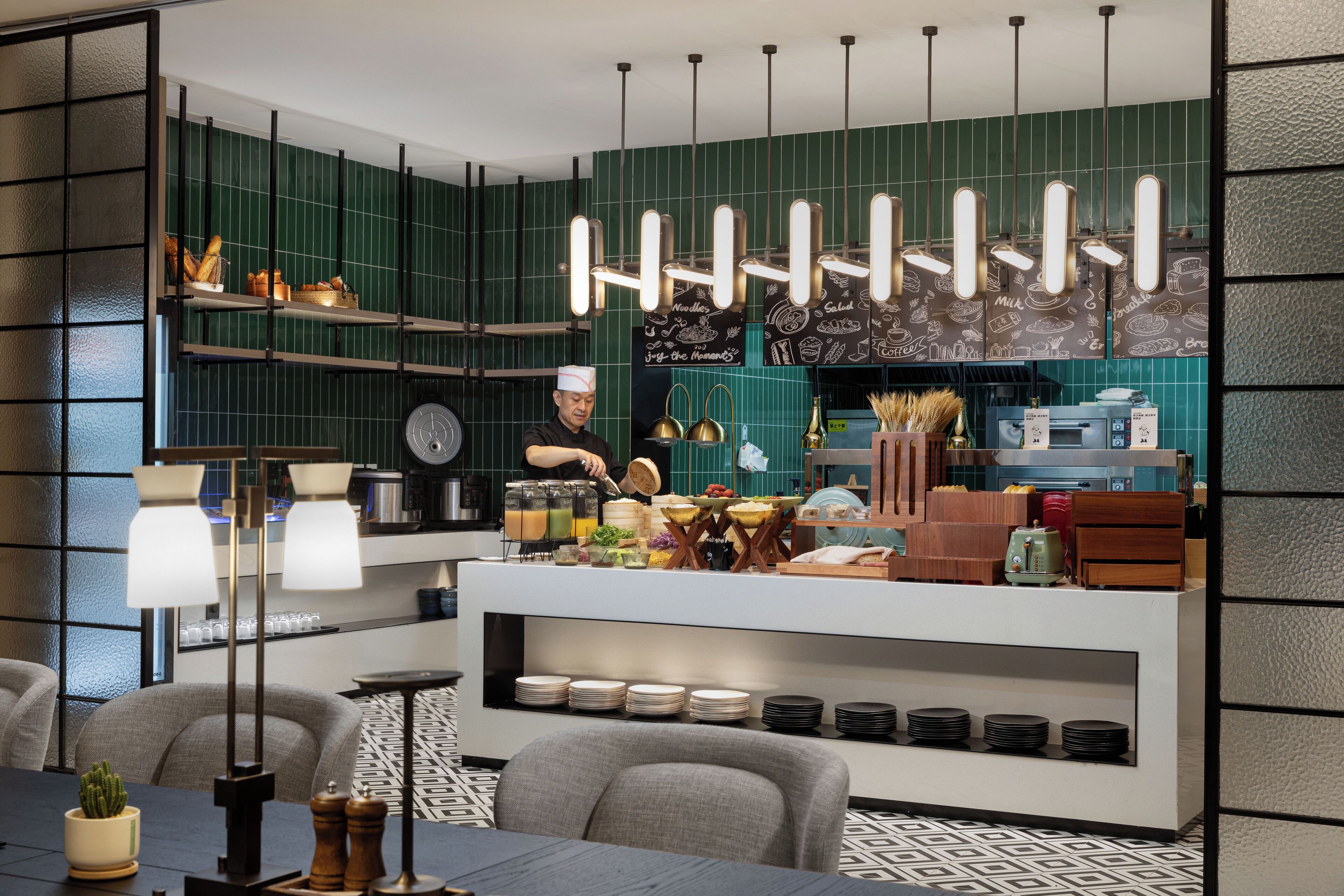 Una cocina moderna con un chef preparando alimentos, un mostrador de buffet con diversos platos, y un comedor con sillas y una mesa.