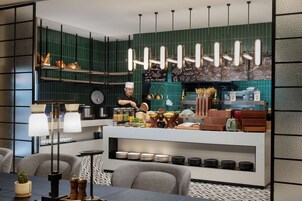 Una cocina moderna con un chef preparando alimentos, un mostrador de buffet con diversos platos, y un comedor con sillas y una mesa.