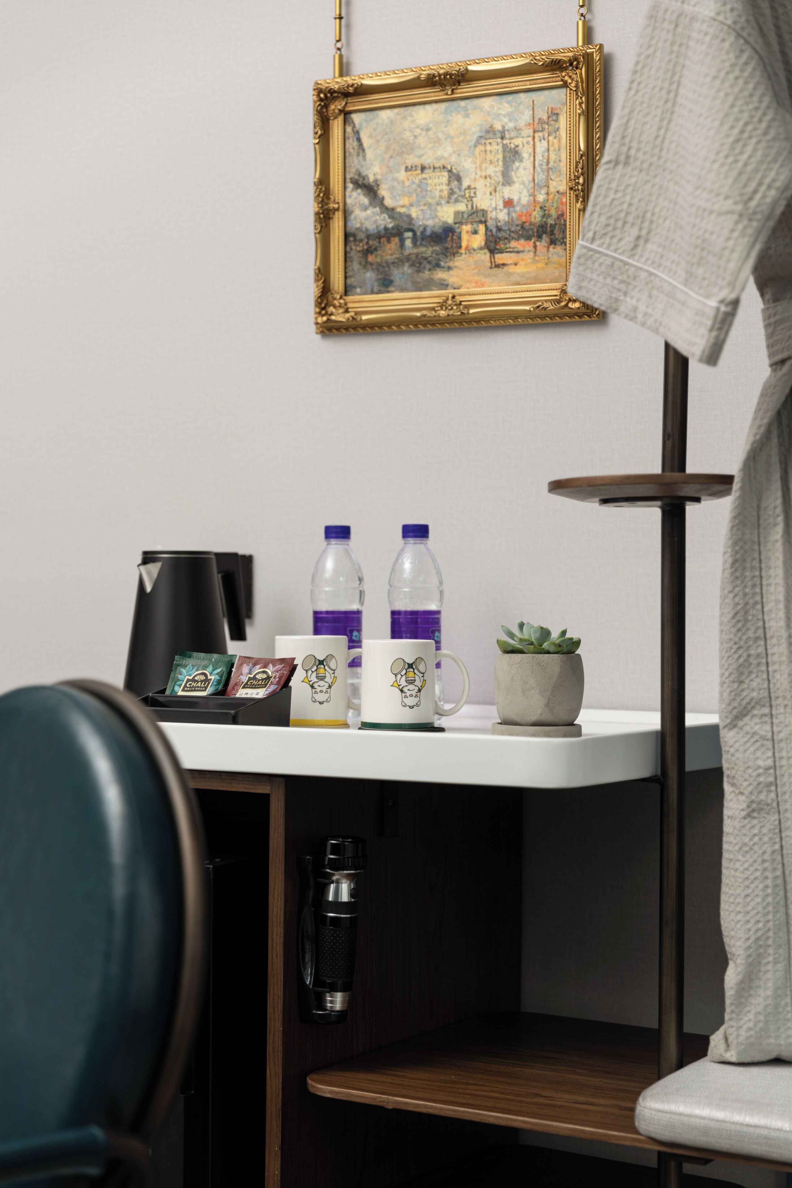 Habitación de hotel con escritorio, un cuadro enmarcado, cafetera, dos botellas de agua, taza y una planta pequeña.